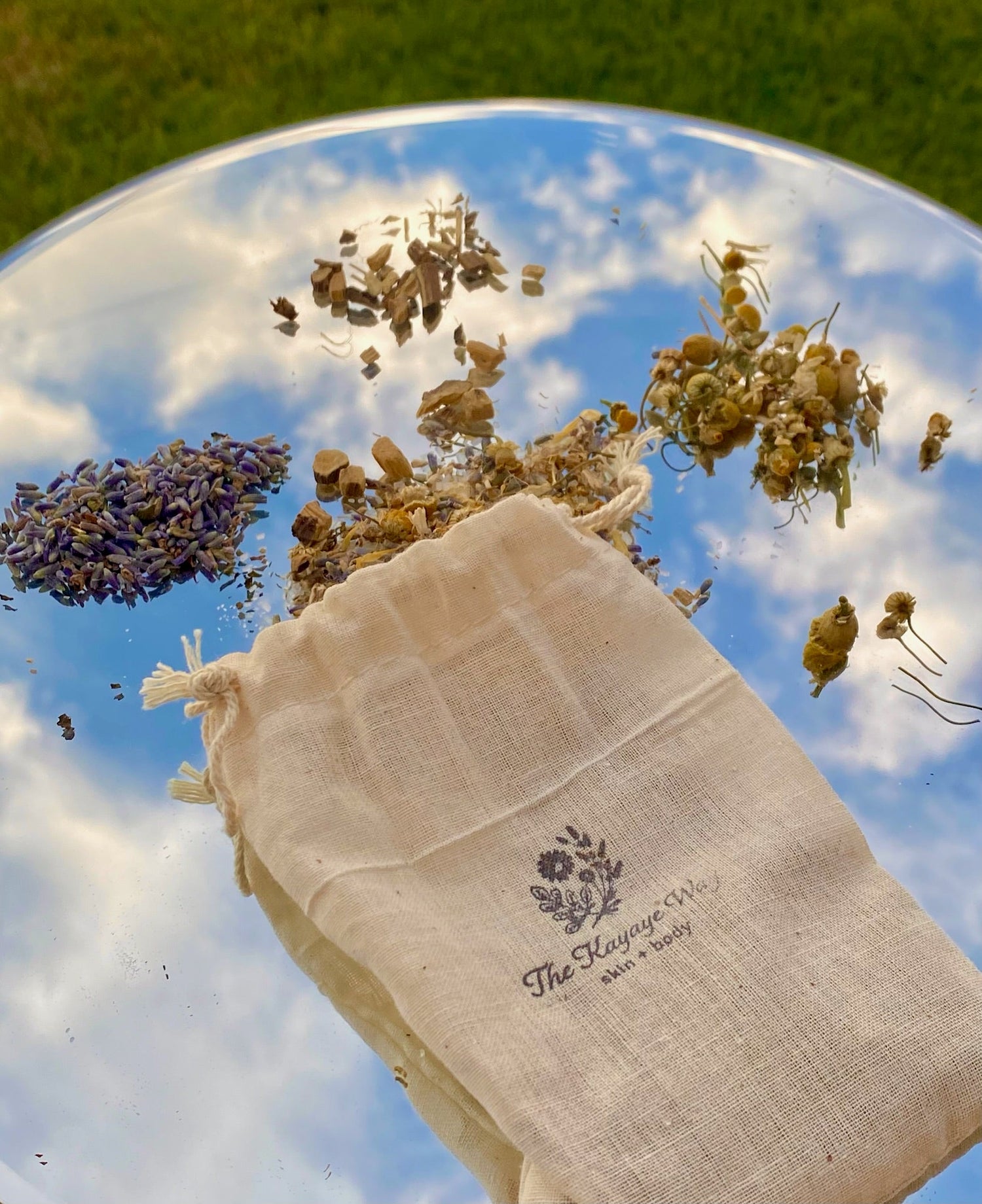 Muslin tea bag sitting on a mirror with various herbs scattered around and a reflection of the blue sky